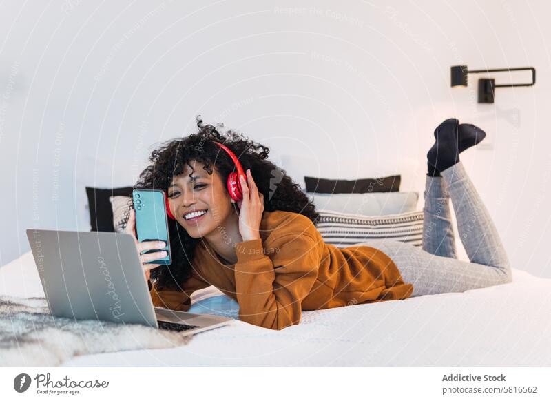 Cheerful black female with laptop and smartphone woman headphones using music netbook rest relax chill portrait african american bed connection browsing mobile