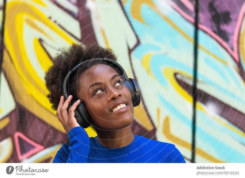 Positive ethnic woman enjoying music in city headphones listen graffiti wall cool millennial urban female black african american afro hairstyle cheerful happy
