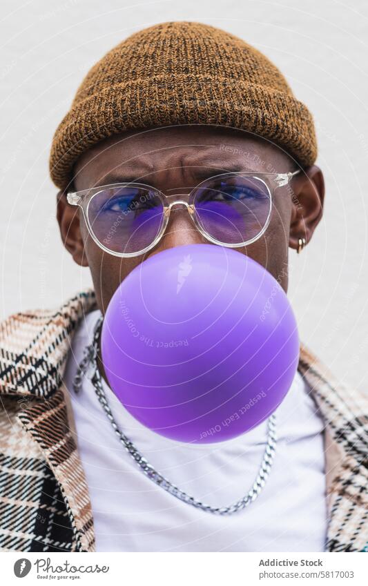 Trendy ethnic man blowing balloon style urban street casual young city modern male black african american hat glasses fashion outfit vivid hipster vibrant wall