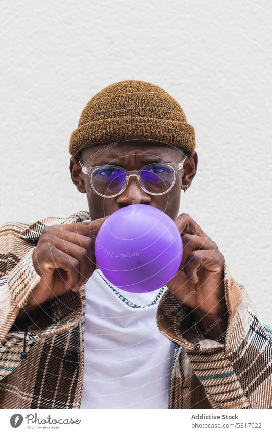 Trendy ethnic man blowing balloon style urban street casual young city modern male black african american hat glasses fashion outfit vivid hipster vibrant wall
