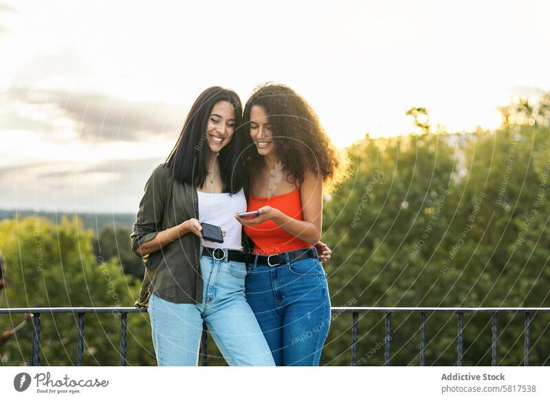two young women smiling and using her mobiles on a terrace. with the sunset people smartphone technology together caucasian girl communication beautiful woman