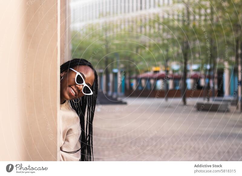 Cheerful ethnic woman peeking from behind column street smile city style fun building young female happy black african american cheerful trendy urban outfit