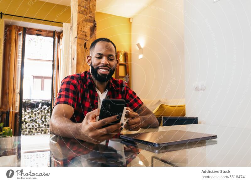 African american man using smartphone while having coffee at home laptop telework computer african american internet freelance remote online black modern