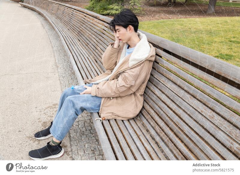 Young man feeling unwell on a park bench, hypoglycemia theme young asian sitting discomfort head hand water bottle jeans jacket outdoor day health sickness pain
