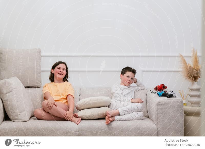 Siblings lounging on sofa at home with toys child sibling girl boy laughing looking at camera pensive looking away decor sitting lounge beige cushion joy