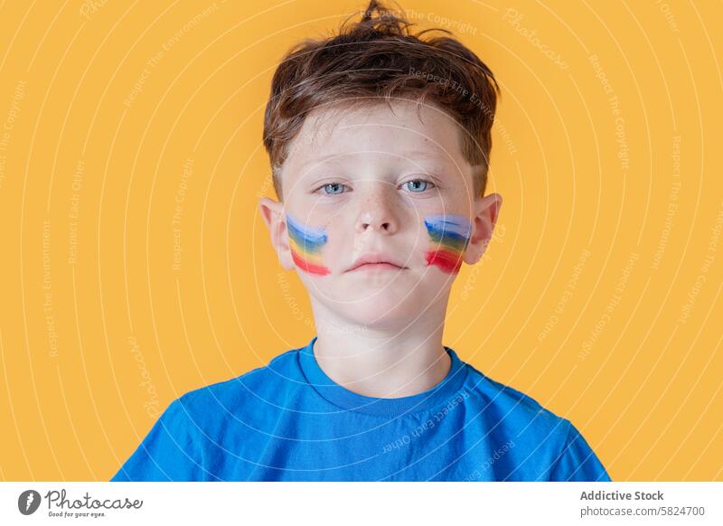Young boy with colorful face paint on a yellow background portrait child serious blue shirt young solemn expression eye contact looking standing vibrant bold