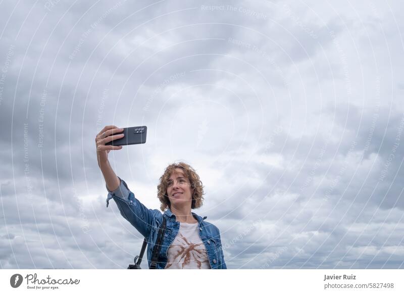 a smiling middle aged woman in casual clothes in her fifties takes a selfie with her mobile phone against a cloudy sky, copy space fourteen 50 40 taking