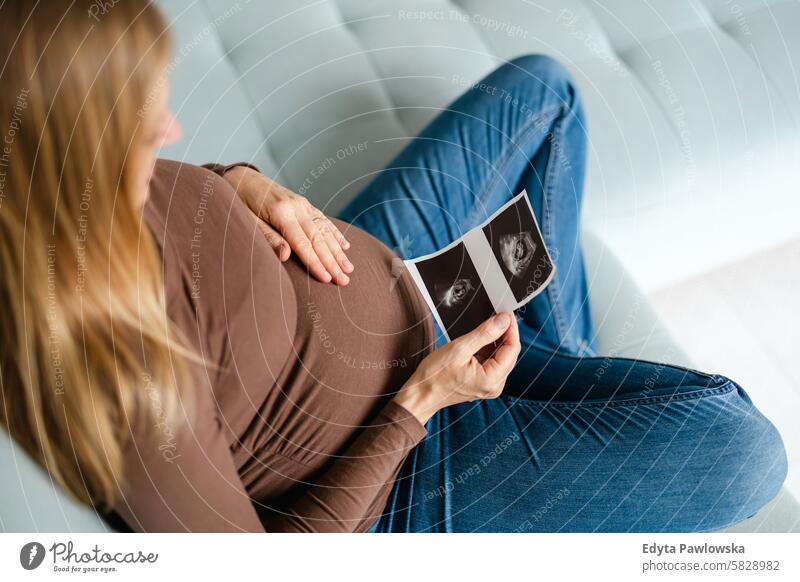 Pregnant woman sitting on sofa at home, looking at the ultrasound scan photo of her baby pregnancy pregnant adult anticipation awaiting belly birth body care
