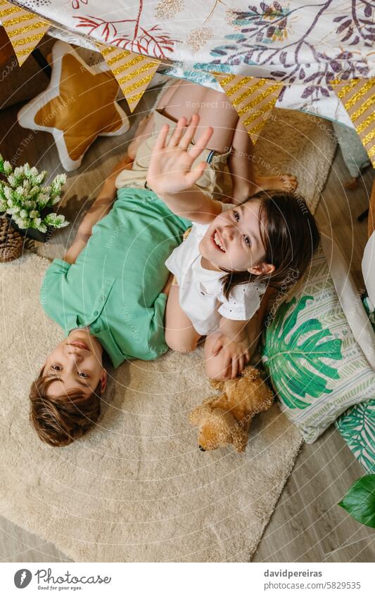Happy little girl raising hand up and smiling boy looking while resting lying over rug in cozy tent. happy smile palm camera looking at camera carpet children