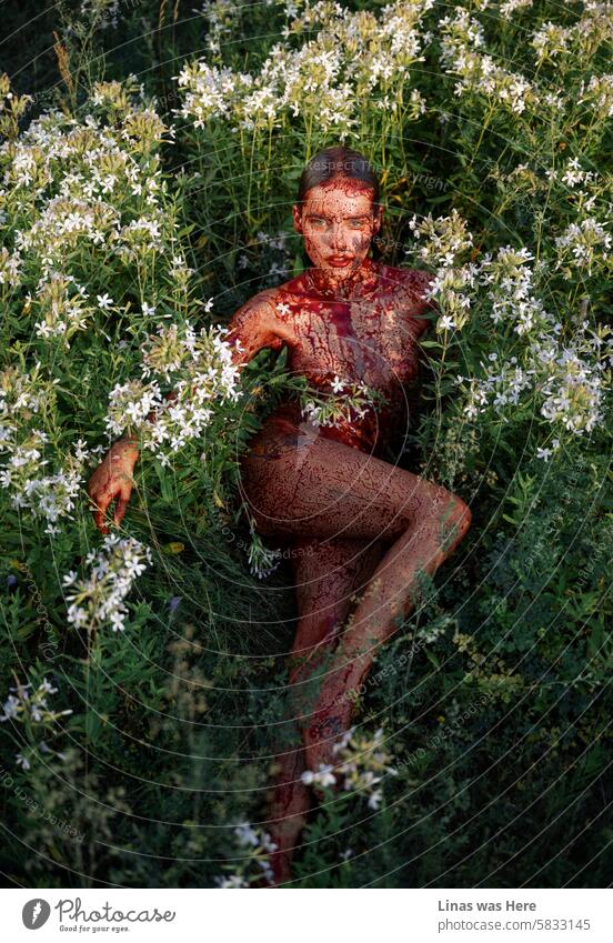 These green fields with white flowers are being occupied by a naked girl covered in fake blood. Like a true satan worshiper, she is feeling just fine in her nude skin. A pretty woman is looking just fine like a devil.