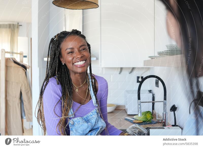 Young woman laughing in a modern kitchen setting cheerful bright braided hair denim overalls lavender shirt joy indoor happiness casual style fashionable