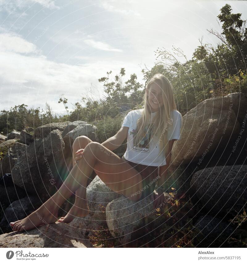 Young, blonde, tall woman with long legs sits barefoot on large stones on the beach and smiles into the camera - analog backlit shot Woman Girl Large 18 years