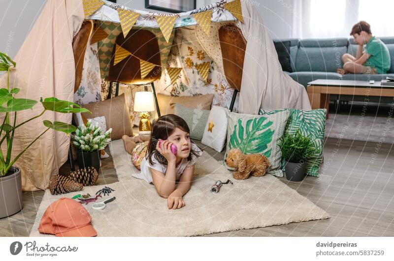 Little girl holding walkie-talkie lying over rug in shelter tent while talk with boy sitting on sofa talking playing carpet couch children teepee cozy vacation