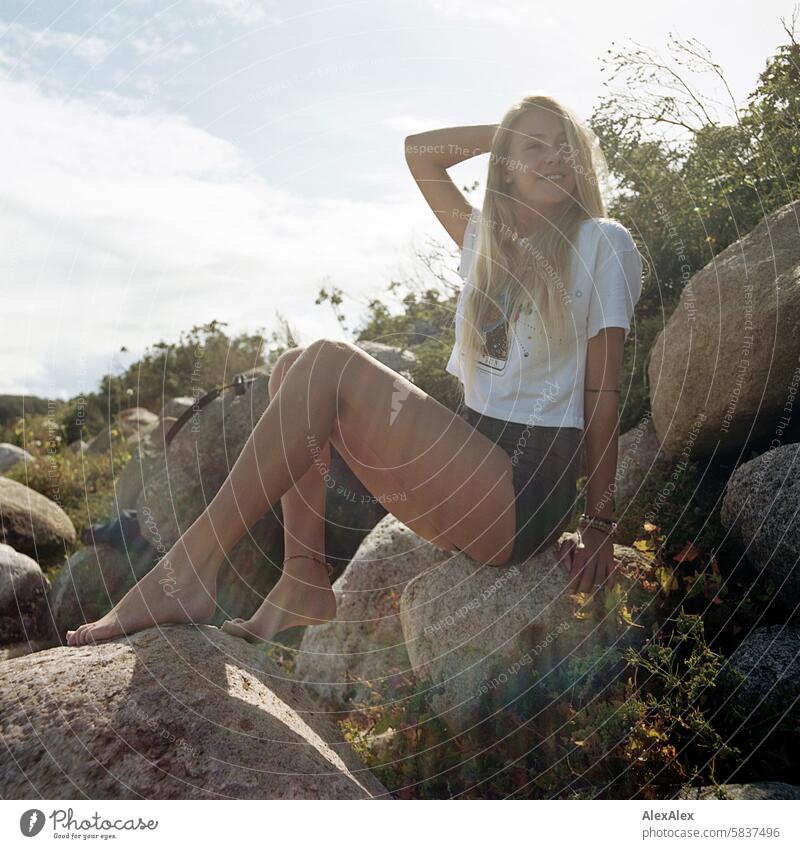 Young, blonde, tall woman with long legs sits barefoot on large stones on the beach and smiles - analog backlit shot Woman Girl Large 18 years Long-haired