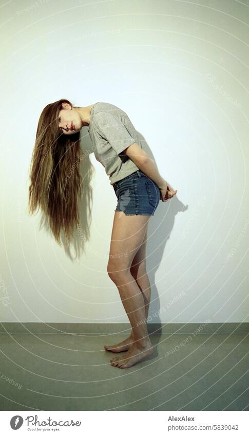 Young, dark-blonde, tall woman stands barefoot in front of a white wall and looks sideways into the camera, her long hair partially covering her face - analog 35mm photo