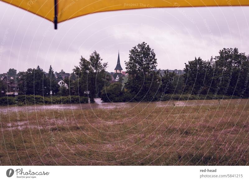 Flood, flooded meadow in the foreground, trees and village with church tower in the background, yellow umbrella of the photographer visible at the top of the picture