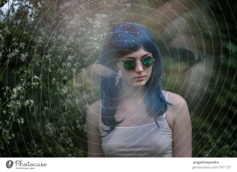 Woman with blue hair in front of bush with white flowers, she wears sunglasses with inscription "love". Wig 70s Love psychedelic Summer petals