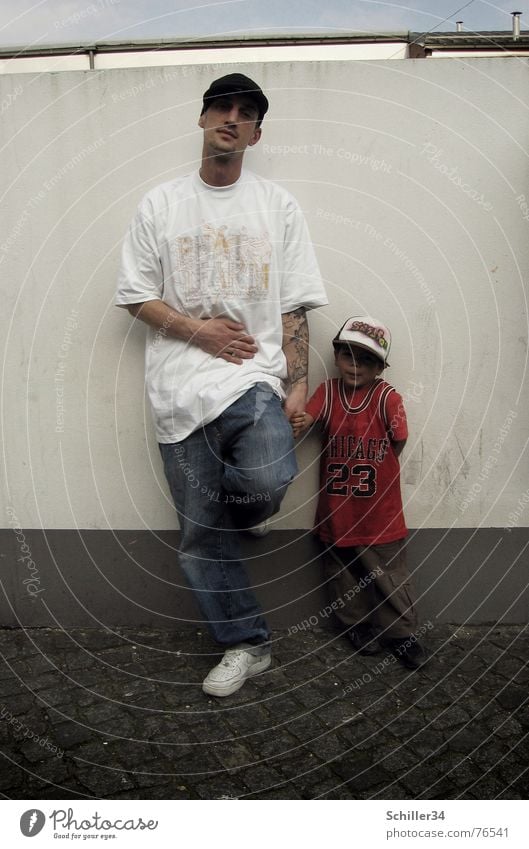 as the father so the... Man Child Father Son Wall (building) Wall (barrier) White Gray Brown T-shirt Red Baseball cap Cap Jersey Hip-hop Sneakers Pants
