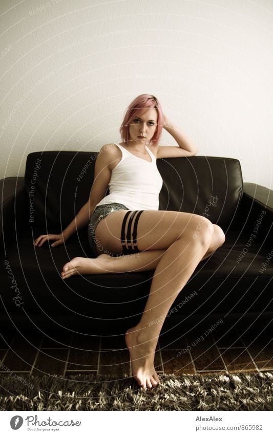 Young, long-legged, slim woman sits barefoot and slanted on a couch. She is barefoot and has three garters on her right leg and pink hair Living room Sofa